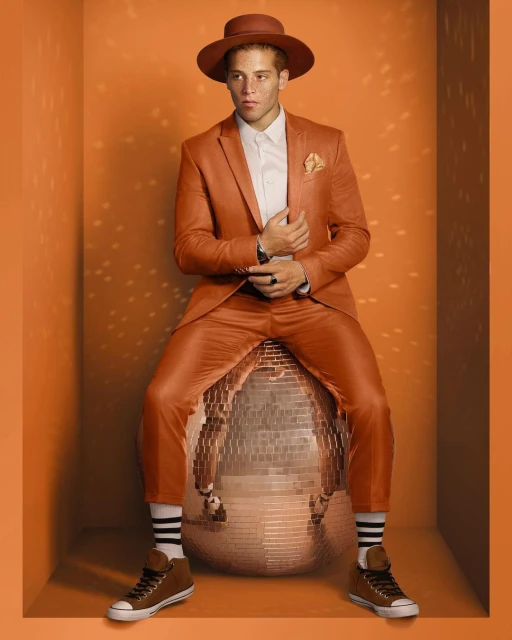 Man in orange suit sitting on disco ball in orange photo studio. He wears a matching hat and brown sneakers with striped socks.